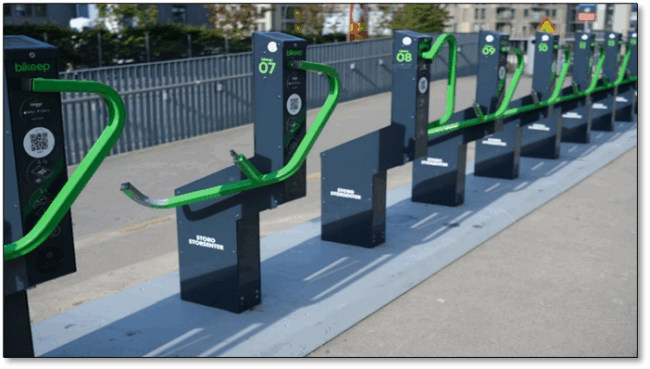 Foto som viser sykkelstativer med integrert l&aring;s ved et kj&oslash;pesenter i Oslo. Stativene er utstyrt med b&oslash;yle og QR-kode. Disse kan gi &oslash;kt sikkerhet, s&aelig;rlig blant syklende som ikke har sikker l&aring;s selv.