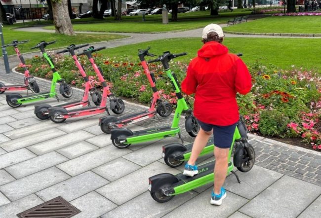 En person med ryggen til og foten p&aring; gr&oslash;nn elsparkesykkel. En rekke av elsparkesykler st&aring;r til venstre for personen.