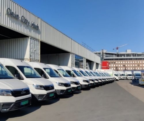 Et bilde som viser elektriske varebiler og lastebiler foran en konstruksjon av sjøkontainere skiltet Oslo City Hub