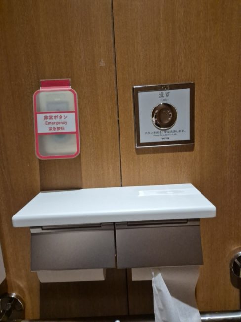 bildet viser en toalettrull-holder som har innebygget hylle p&aring; oversiden hvor det er mulig &aring; legge fra seg utstyr. Det er ogs&aring; bilde av en alarmknapp for assistanse som er montert like ved rullen.