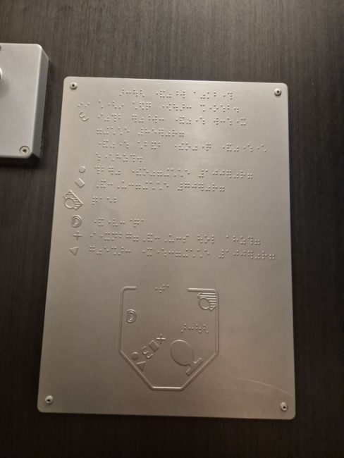 bildet viser et taktilt skilt p&aring; et tog-toalett. Skiltet gj&oslash;r det mulig &aring; f&aring; oversikt over rommet uten &aring; m&aring;tte f&oslash;le seg fram med hendene da b&aring;de vask, toalett, d&oslash;r og toalettruller er markert med taktile symboler.