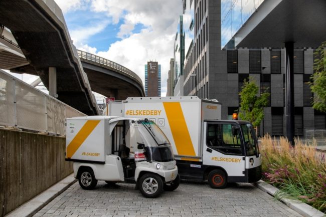 foto av en firehjuls overbygget moped med lasteskap (Paxster) og et lite lastekjøretøy med skap og en tilhenger. Begge kjøretøyene er merket med Elskedeby sine farger og logo.