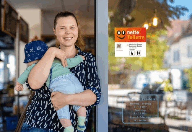 Bildet viser en dame og en baby som st&aring;r i en d&oslash;r&aring;pning p&aring; en kafe. Det henger et skilt p&aring; kafeen hvor det st&aring;r &laquo;Nette toilette&raquo; for &aring; vise at kafeen er med i ordningen.
