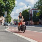 Gravid kvinne som sykler en tohjuls lastesykkel med lasteplan foran. I lasteplanet sitter en eldre dame og et barn. Bilde er tatt på sommeren i Strømsveien i Oslo. De sykler på separat sykkelinfrastruktur med rød asfalt.