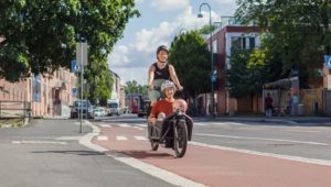 Gravid kvinne som sykler en tohjuls lastesykkel med lasteplan foran. I lasteplanet sitter en eldre dame og et barn. Bilde er tatt på sommeren i Strømsveien i Oslo. De sykler på separat sykkelinfrastruktur med rød asfalt.