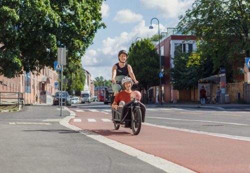 Gravid kvinne som sykler en tohjuls lastesykkel med lasteplan foran. I lasteplanet sitter en eldre dame og et barn. Bilde er tatt på sommeren i Strømsveien i Oslo. De sykler på separat sykkelinfrastruktur med rød asfalt.