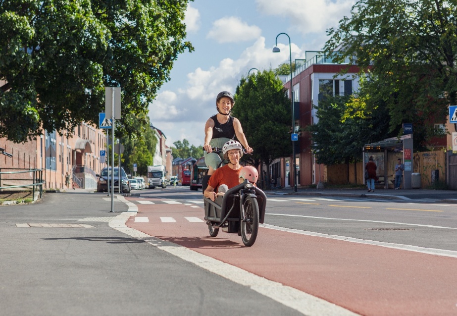 Gravid kvinne som sykler en tohjuls lastesykkel med lasteplan foran. I lasteplanet sitter en eldre dame og et barn. Bilde er tatt p&aring; sommeren i Str&oslash;msveien i Oslo. De sykler p&aring; separat sykkelinfrastruktur med r&oslash;d asfalt.