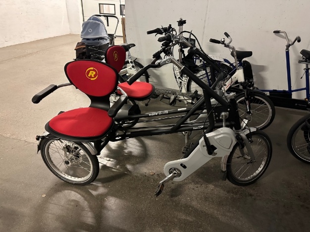 Sykkel for én sjåfør og én syklist, der man sitter ved siden av hverandre. Det ene sete er vendt 90 grader fra styret, som gjør det lettere å stige på sykkelen. Sykkelen er svart og grå med røde setter med rygg- og armstøtte. Bildet er tatt en garasje.