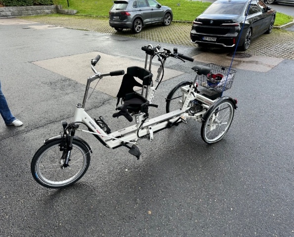 Tandemsykkel med tre hjul, ett foran og to bak. Det er pedaler og bremser til begge syklister. Sete foran har ekstra støtte i ryggen. Det er en kurv bakerst på sykkelen. Bildet er tatt på en parkeringsplass.